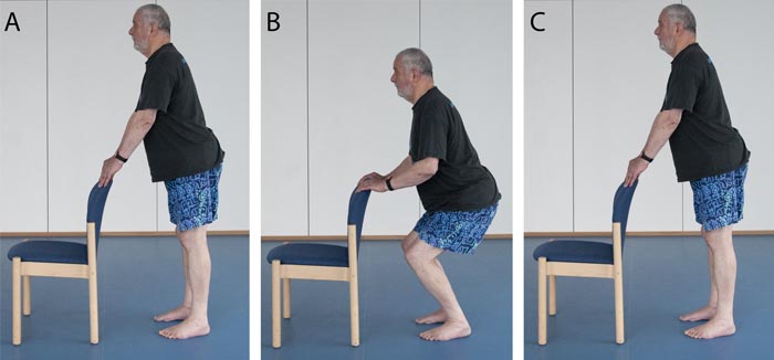 A man holds the back of a chair and lowers himslef into a comfortable squat.