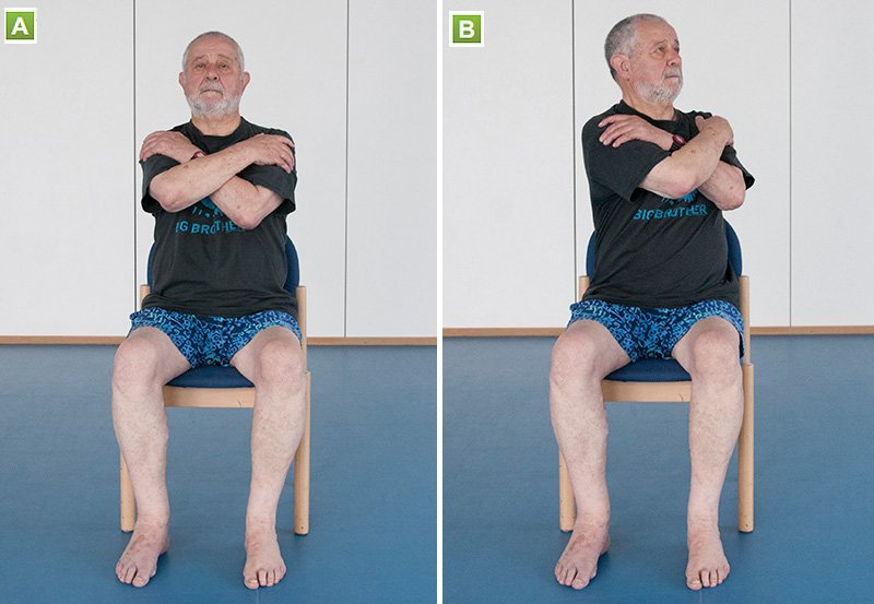 A man sits with his arms crossed holding the oppoiste upper arm, he then twists his upper body, while keeping his bottom half straight